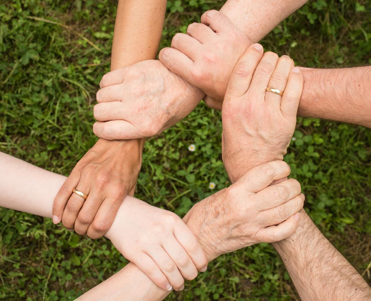 Six hands of different people clasped together in a circle over green grass